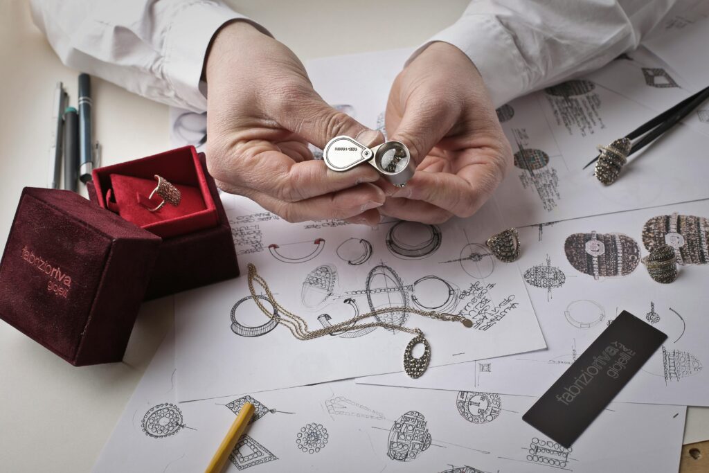 pexels-photo-1050321-1050321-1 Close-up of hands inspecting a diamond ring with a loupe on a workbench filled with jewelry sketches.
